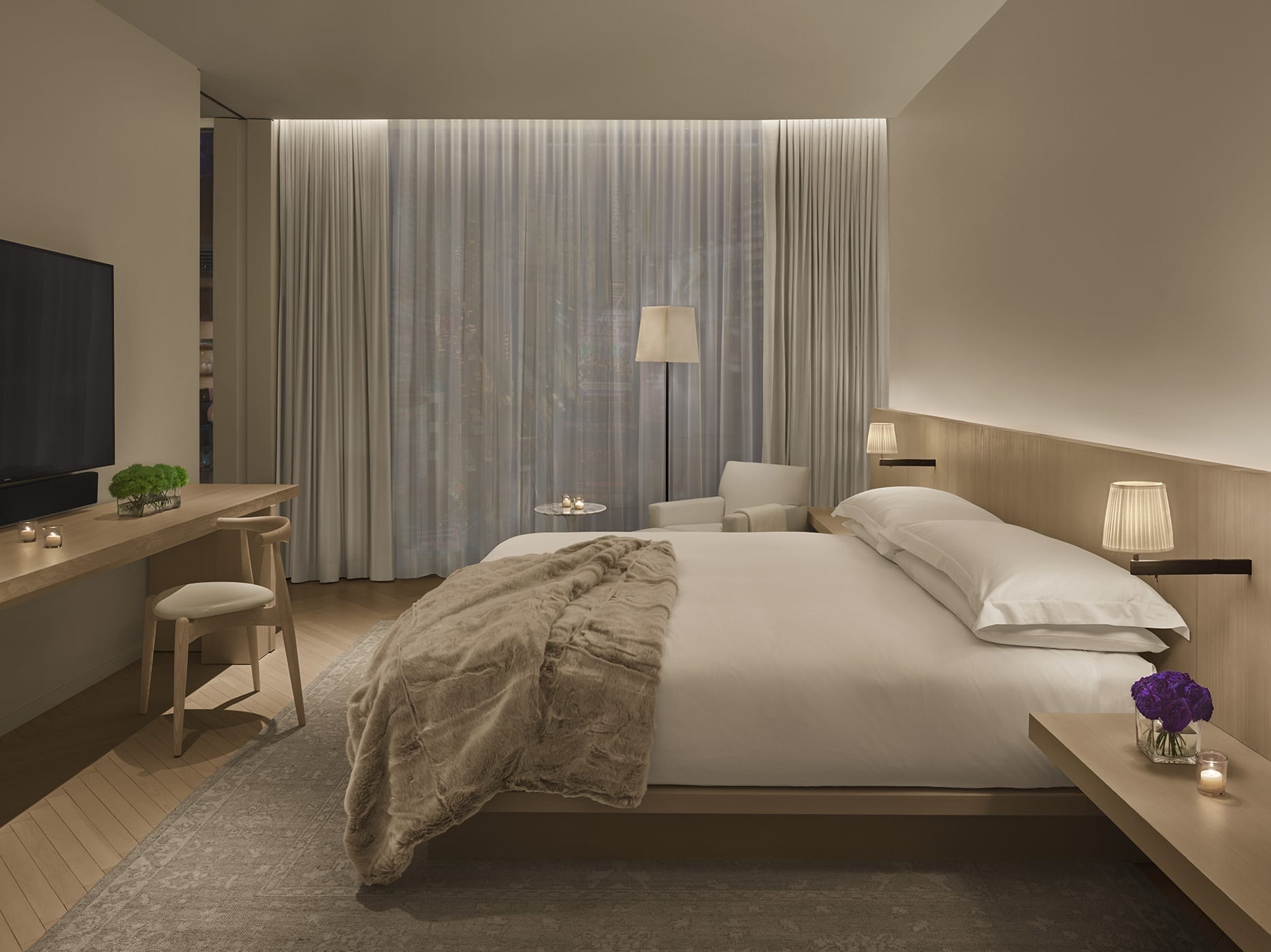 Serene bedroom at the Times Square Edition, with a king bed, pale oak and cream decor, a faux-fur throw and Times Square's neon glowing through sheer curtains