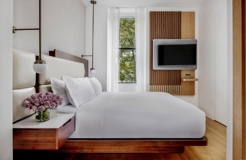 Bright bedroom at the Bryant Park Hotel in Midtown Manhattan, with a king bed, warm wood furnishings, a vase of lilac roses and a window onto leafy treetops